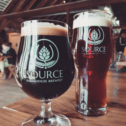 Two glasses of beer on a wooden table, one dark and one amber, both featuring the Source Farmhouse Brewery logo.