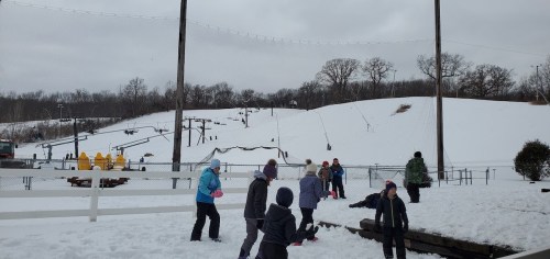 Children playing in the snow at a winter park, with ski lifts and trees in the background under a cloudy sky.
