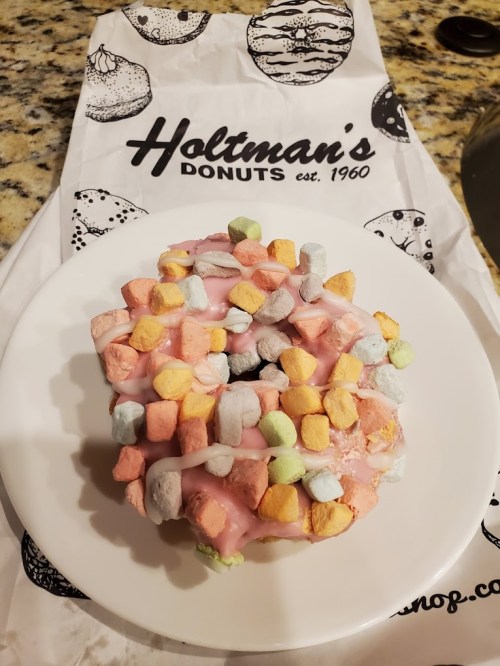 A colorful donut topped with pastel marshmallows on a white plate, resting on a branded donut bag.
