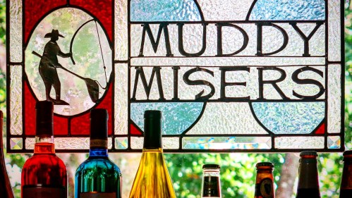Stained glass window with "Muddy Misers" and colorful bottles of wine and beer below it.