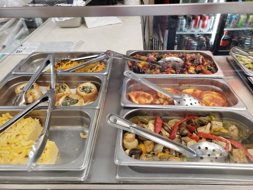 A display of various dishes in metal trays, including pasta, vegetables, and baked items, with serving utensils.