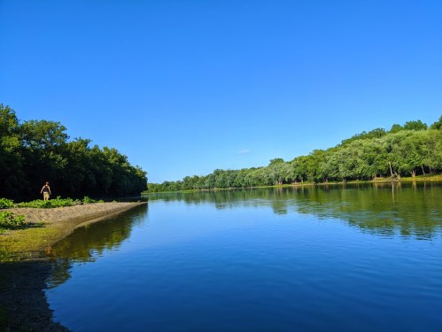 A serene river scene with clear blue skies, lush green trees lining the banks, and a person standing on the shore.