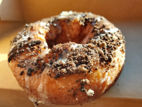 A close-up of a glazed donut topped with crushed cookies and a drizzle of icing, resting in a cardboard box.