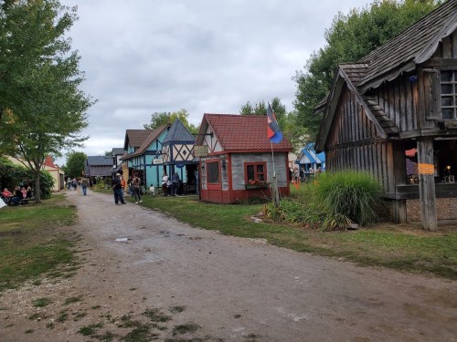 A quaint village scene with colorful houses, a dirt path, and people enjoying the outdoors under a cloudy sky.