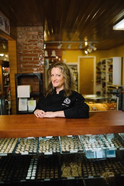 A smiling woman in a black chef's coat stands behind a counter filled with assorted chocolates and confections.