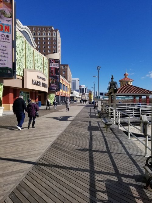 A sunny boardwalk scene with people walking, shops on one side, and a gazebo by the water on the other.