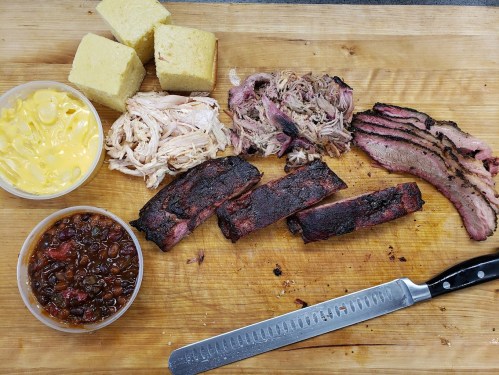 A wooden board with sliced brisket, pulled pork, chicken, cornbread, and baked beans, alongside a knife.