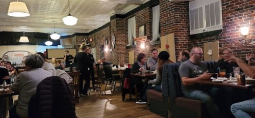 A cozy restaurant interior with brick walls, diners at tables, and a server attending to guests. Warm, inviting atmosphere.