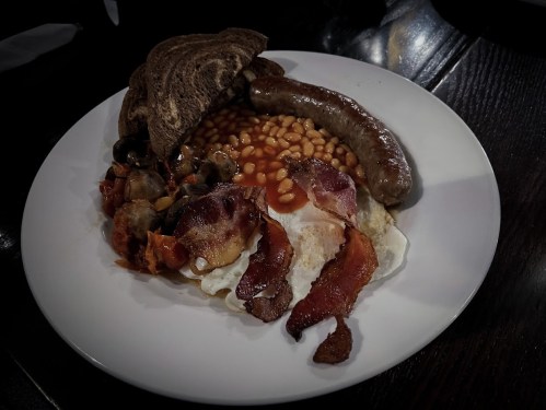 A plate of breakfast featuring eggs, bacon, sausage, baked beans, mushrooms, and toast.