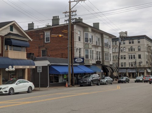 A street view featuring buildings with shops, parked cars, and a cloudy sky.