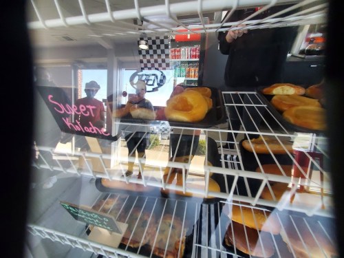 A display case filled with pastries, with a sign reading "Sweet Killed" and reflections of people in the background.