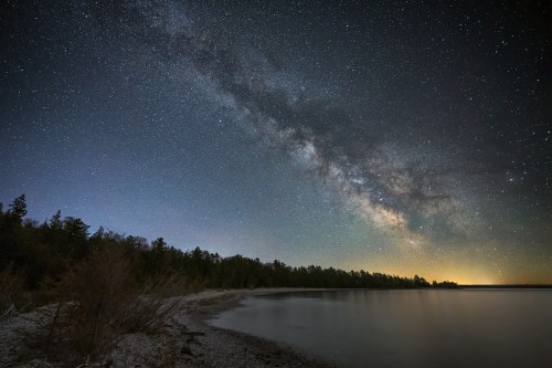 A serene lakeside view under a starry night sky, featuring the Milky Way and a calm water reflection.