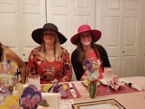 Two women wearing large hats sit at a beautifully set dining table, smiling at the camera.