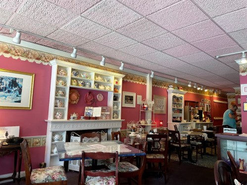 Cozy café interior with pink walls, vintage decor, and wooden tables, featuring shelves of teacups and a warm atmosphere.