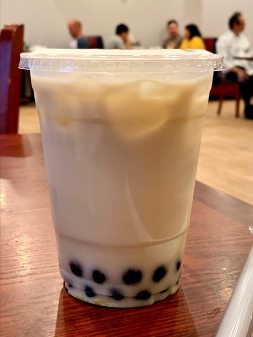 A clear cup filled with creamy beverage and black tapioca pearls, placed on a wooden table in a cafe setting.