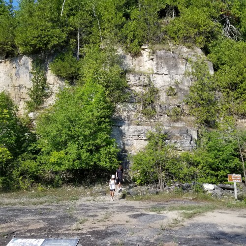 Two people walk towards a rocky cliff surrounded by lush green trees and vegetation on a sunny day.