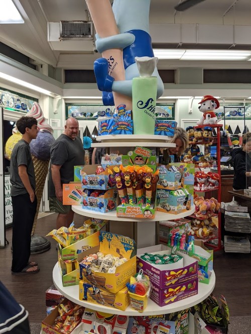 A colorful display of snacks and toys in a shop, with customers browsing in the background and a large ice cream figure above.