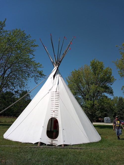 A white teepee with a door and poles on top, set in a grassy area with trees and a clear blue sky in the background.