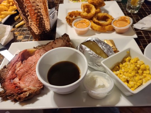 A plate featuring prime rib with au jus, a baked potato, corn, and onion rings in the background.