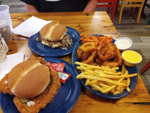 Two plates of food featuring sandwiches, fries, sweet potato fries, onion rings, and dipping sauces on a wooden table.