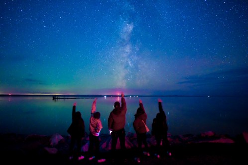 Silhouetted figures point towards a starry sky and the Milky Way over a calm lake at night.