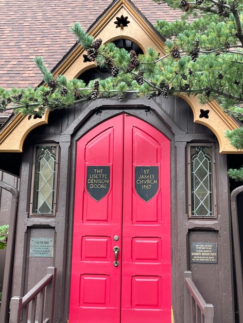 Red double doors of St. James Church, framed by decorative woodwork and surrounded by pine branches.