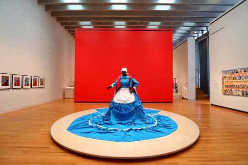 A figure in a blue dress stands on a circular platform in an art gallery, with a red wall and framed artworks in the background.