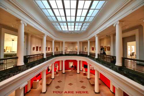 A spacious art gallery with columns, skylights, and red walls, featuring the text "YOU ARE HERE" on the floor.