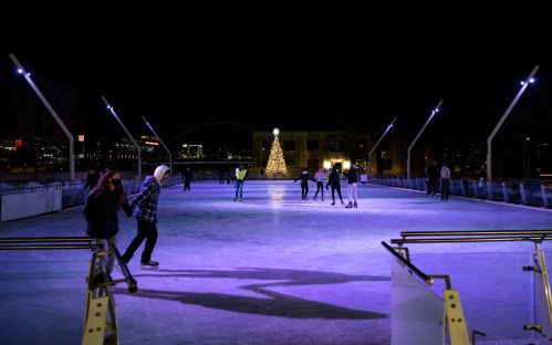 People ice skating at night on a rink, with a Christmas tree illuminated in the background.