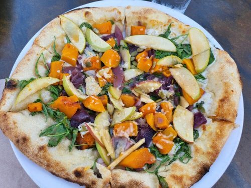 A pizza topped with butternut squash, apples, greens, and drizzled sauce on a golden crust, served on a white plate.