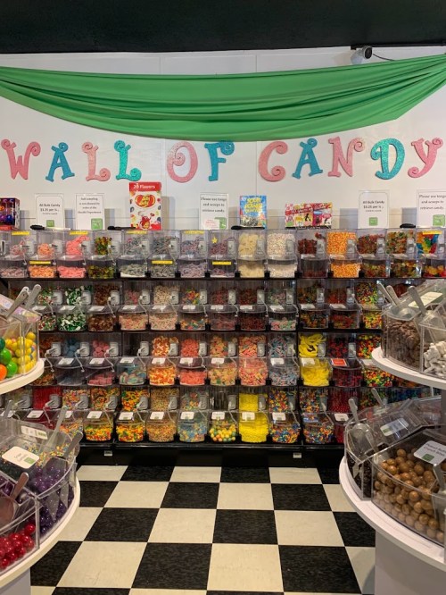 A colorful candy wall filled with various jars of sweets, with a green banner reading "WALL OF CANDY" above.