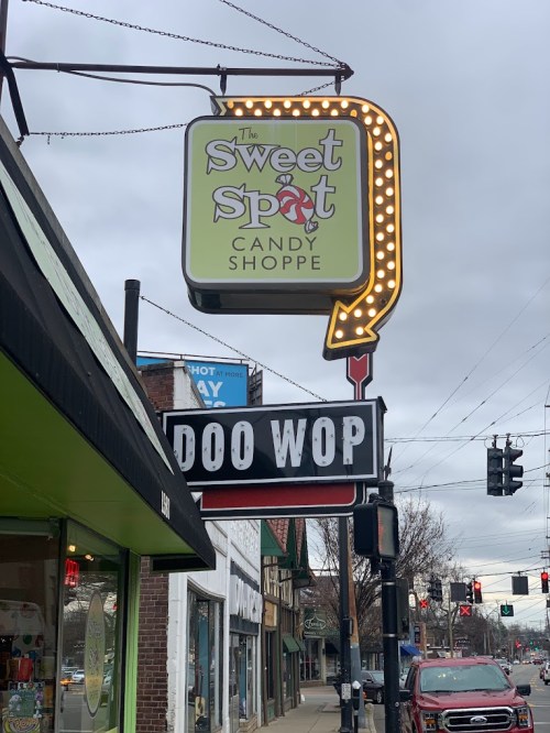 Sign for "The Sweet Spot Candy Shoppe" with a retro "Doo Wop" sign below, set against a cloudy sky.