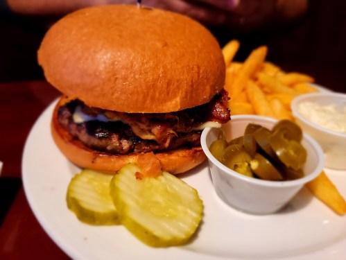 A juicy burger with bacon and cheese, served with pickles, jalapeños, and a side of golden fries on a white plate.