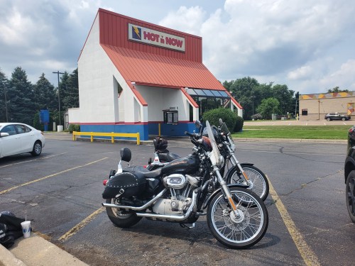 Two motorcycles parked in front of a "Hot 'n Now" fast food restaurant on a sunny day.