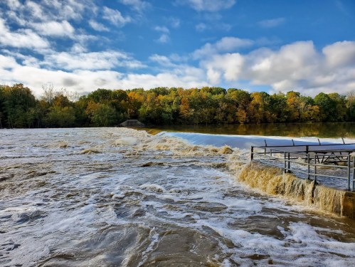 A river overflowing a dam, with foamy water cascading down and colorful autumn trees in the background under a blue sky.