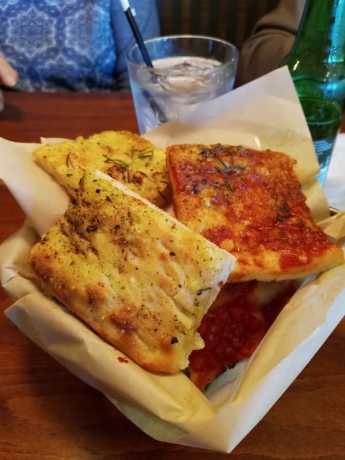 A basket of assorted bread, including garlic bread and pizza slices, with a glass of water and a green bottle in the background.