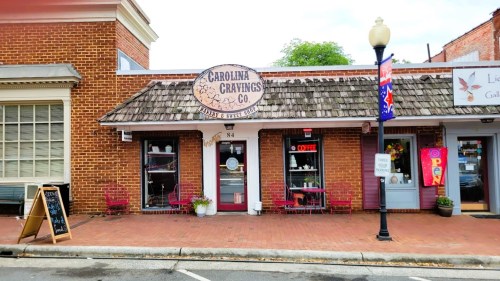 A quaint storefront with a sign reading "Carolina Cravings Co." and outdoor seating in a charming brick-lined street.