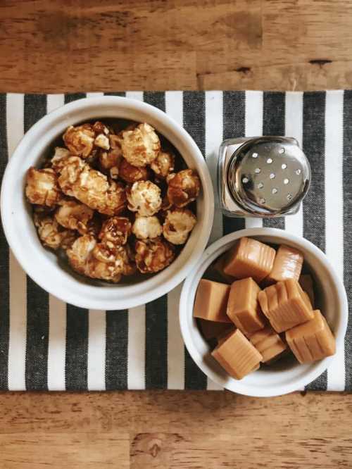 A bowl of caramel popcorn, a bowl of caramel squares, and a salt shaker on a striped cloth.