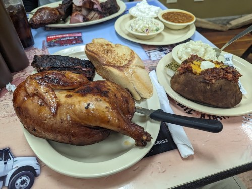 A plate featuring a roasted chicken, toast, a baked potato with toppings, and sides of coleslaw and beans.