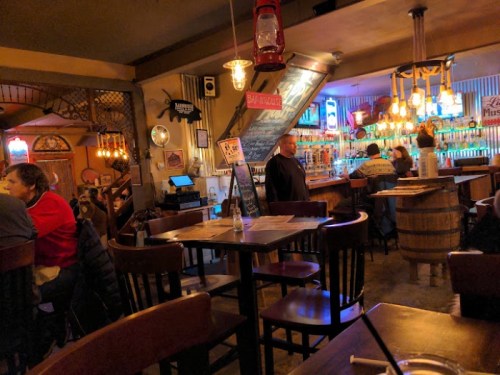 A cozy bar interior with wooden tables, warm lighting, and patrons enjoying drinks at the counter.