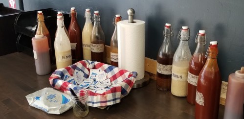 A table with various sauce bottles, napkins, and a jar, set against a blue wall.