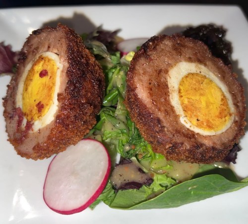 A close-up of two sliced scotch eggs on a bed of mixed greens, garnished with radishes and a dark sauce.