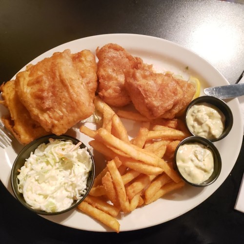 A plate of fried fish, golden fries, coleslaw, and two small bowls of dipping sauce.