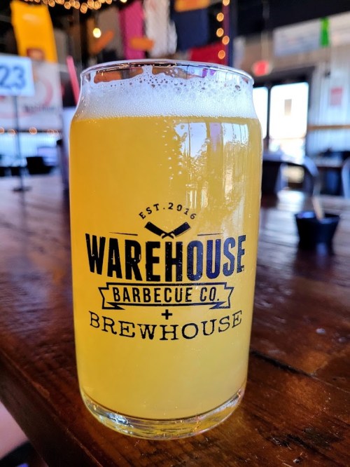 A glass of light beer sits on a wooden table in a lively barbecue restaurant.