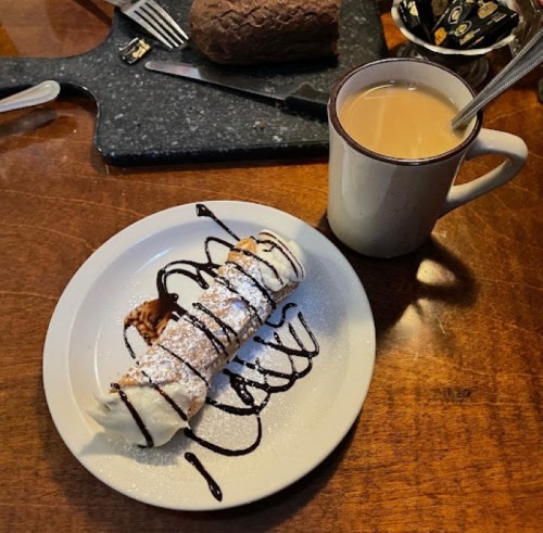 A plate with a cannoli drizzled in chocolate and powdered sugar, next to a cup of coffee on a wooden table.