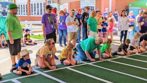 A group of people, including children and adults, kneel on a green track at an outdoor event, preparing for a race.