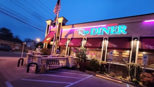 A brightly lit diner at dusk, featuring neon signage and outdoor seating, with an American flag flying.