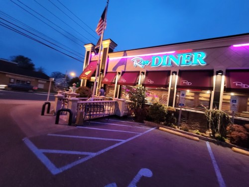 A brightly lit diner at dusk, featuring neon signage and an American flag, with parked cars in the foreground.
