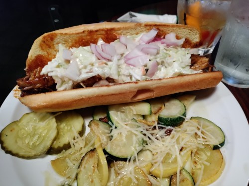 A large sandwich filled with pulled pork, coleslaw, and onions, served with sautéed zucchini and pickles on the side.