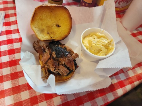 A pulled pork sandwich on a bun with a side of creamy macaroni and cheese, served on a red and white checkered tablecloth.